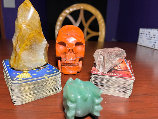 Two Tarot Decks Side By Side one with golden healer crystal flame a Red Jasper Skull Carving Separates the two with the other deck having a Lemurian Seed Crystal on top of it. 